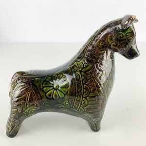 May include: A ceramic horse figurine in shades of brown and green. The horse has a glossy finish and is decorated with floral and scroll patterns. The figurine is standing on four legs and has a curved neck and head.