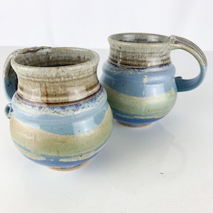 May include: Two handmade ceramic mugs with blue, green, and brown glaze. The mugs have unique, asymmetrical handles.