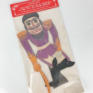 May include: A vintage paper doll of the Nutcracker from the "Jumping Jacks" series. The doll is dressed in a purple and gold uniform with a black hat and a long, orange stick. The doll is in the style of a traditional Nutcracker.