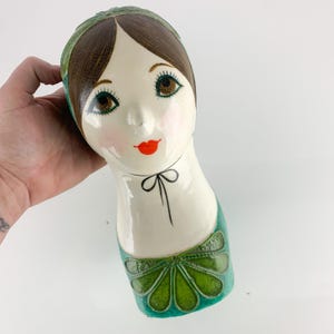 May include: A vintage ceramic head vase with a stylized face. The face has brown hair, brown eyes, and red lips. The vase is painted with green accents on the head covering and neckline. The vase is held by a hand.
