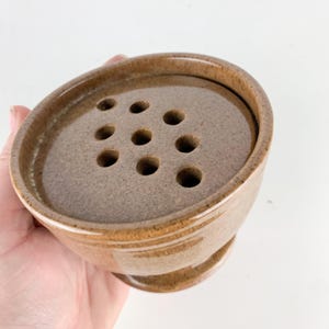 May include: A round, brown ceramic flower frog with a speckled glaze finish. The top surface features nine evenly spaced circular holes arranged in a grid pattern, designed to hold individual flower stems upright within the bowl-shaped base.