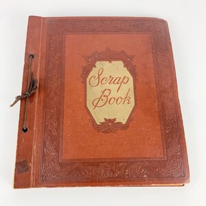May include: A vintage scrapbook with a reddish-brown cover. The cover features embossed floral designs and a gold-colored label with the words "Scrap Book". A brown string ties the book closed. The scrapbook is a nostalgic item for preserving memories.