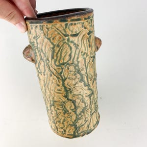 May include: A handmade ceramic vase with a textured, tree-like design. The vase features a beige base with green and brown accents, and two small handles. The top rim is dark brown.