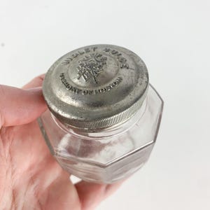 May include: A vintage glass jar with a silver lid. The lid has the text "Wrigley's Dulce" and "Factory of Boston" embossed on it.
