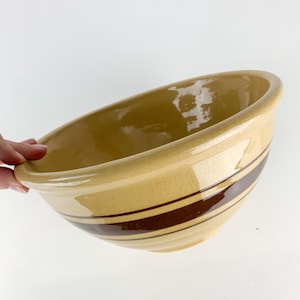 May include: A large, round ceramic mixing bowl with a glossy, light tan finish and a distinct crackle glaze texture. The exterior features two prominent, horizontal dark brown bands encircling the middle section of the bowl.