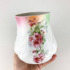 May include: White ceramic vase with a floral rose design. The vase has a curved shape and is decorated with red roses and green leaves. The top of the vase has a gradient of pink, orange, and green.