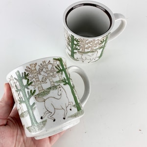 May include: Two ceramic mugs featuring a white base with a speckled finish. The exterior is decorated with a repeating pattern of green and brown stylized trees and white horses. Each mug has a curved handle and a dark brown interior rim.