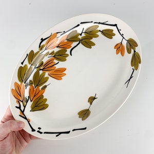 May include: A white oval serving platter with a black and brown floral design. The design features stylized branches with orange and green leaves.