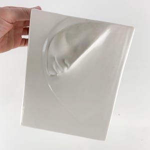 May include: A white ceramic wall hanging with a unique, abstract design. The piece features a raised, curved form on a rectangular base. The artwork is held by a hand, showcasing its shape and texture. The overall aesthetic is minimalist and modern.
