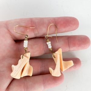 May include: A pair of earrings with gold-colored hooks. Each earring features a cluster of small white beads and a larger, abstract, peach-colored pendant. The earrings are displayed against a neutral background.