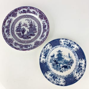 May include: Two decorative plates with intricate designs. One plate features a purple and white pattern with a pagoda scene. The other plate has a blue and white design with a similar pagoda and floral motif. Both plates have detailed borders.