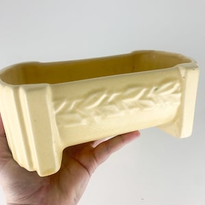 May include: A pale yellow ceramic planter with a raised design of a continuous row of leaves. The planter is rectangular with rounded edges and has a slightly curved bottom.