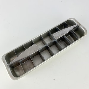 May include: A vintage metal ice cube tray with a rectangular shape and individual square compartments. A metal divider is in the center. The tray is made of a silver-colored metal and is likely from the mid-20th century.