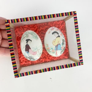 May include: Two hand-painted eggs in a glass display case with a red velvet lining. The eggs feature traditional Chinese artwork of women in colorful clothing.