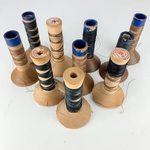 May include: Nine wooden spools of thread, some with blue and red labels, and some with black thread. The spools are arranged on a white surface.