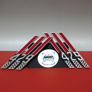 May include: A collection of automotive emblems with a red and white stripe design, featuring the number "429" in white. The emblems have a checkered flag pattern at the base and are displayed on a black stand.