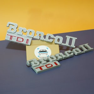 Puede incluir: Emblemas grises de plástico Bronco II TDI. La palabra "Bronco" está en letras grandes, con "TDI" en rojo debajo. El número "II" está al lado de la palabra "Bronco". El fondo es una mezcla de amarillo y azul.