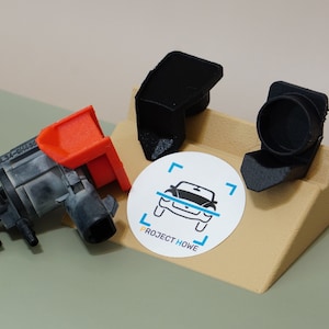 May include: Three automotive components in black, grey, and red are displayed on a tan platform. A white circular sticker with a car graphic and the text "PROJECT HOWE" is also present. The parts appear to be 3D-printed.