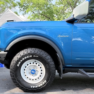 Pode incluir: Um SUV Ford Bronco azul com uma roda branca e um pneu preto. O pneu tem o texto "GRABBER A/T" e "GENERAL" em branco. A palavra "Bronco" é visível na lateral do veículo.