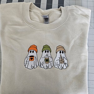May include: A beige sweatshirt with three embroidered ghosts wearing hats and holding beverages. The ghosts are wearing orange, green, and brown hats. The ghosts are holding a cup of coffee, a cup of iced coffee, and a cup of tea.