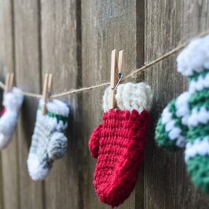 May include: A string of crocheted mitten ornaments in various colours, including red, green, white, and grey, are hung on a wooden fence with twine and clothes pegs.
