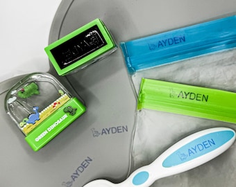 Puede incluir: Un conjunto de cuatro sellos personalizados verdes y azules con el nombre "AYDEN" impreso en ellos. Los sellos están sobre una superficie gris con un sello de dinosaurio verde y una cuchara blanca con mango azul.