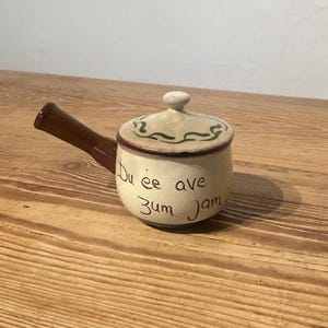 May include: A small, off-white ceramic pot with a lid and a brown handle. The lid is decorated with a green and brown design. The pot has the words "Du ee ave zum jam" written on it in black ink.