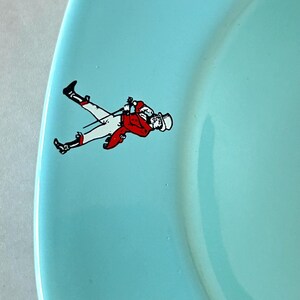 May include: A light blue plate with a decorative illustration of a person in a red jacket, white pants, and a top hat. The person is holding a riding crop. The plate has a curved edge.