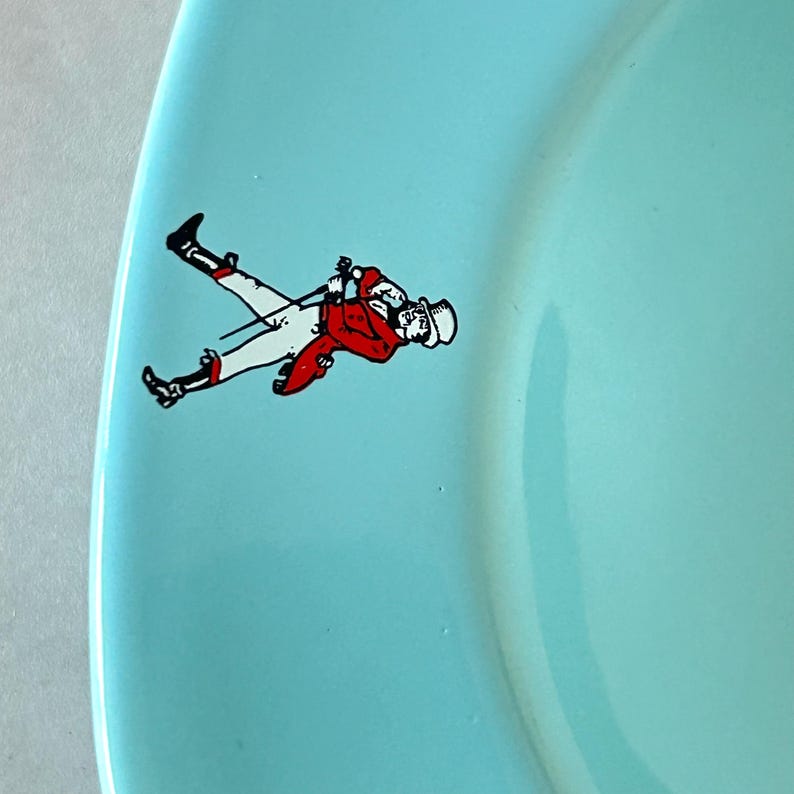 May include: A light blue plate with a decorative illustration of a person in a red jacket, white pants, and a top hat. The person is holding a riding crop. The plate has a curved edge.