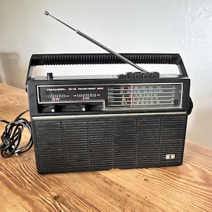 May include: A black Realistic DX-55 portable radio featuring an extendable metal aerial, a top-mounted carrying handle, and a front-facing speaker grille. The radio display shows FM, AM, and shortwave frequency bands with manual tuning sliders.