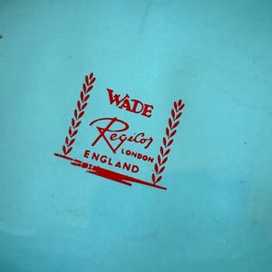 May include: A close-up of a light blue surface with a red logo. The logo reads "Wade Regicor London England" with decorative elements. The text is in a stylized font.
