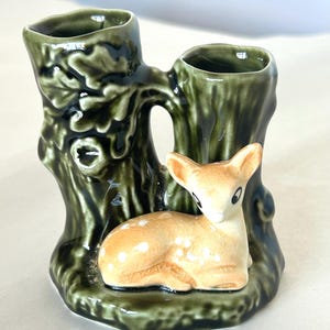 May include: A ceramic vase in the shape of a tree trunk with two openings, painted in shades of green. A small fawn with white spots is nestled at the base of the vase. The fawn is tan with black eyes and ears.