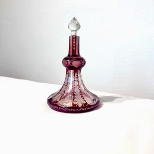 May include: A cranberry red glass decanter featuring a clear glass stopper and intricate etched floral patterns. The vessel has a wide, flared base that tapers into a slender neck, with a bulbous midsection decorated with heart-shaped motifs.