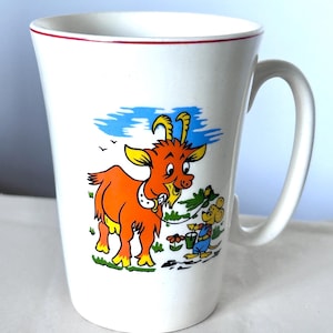 May include: White ceramic mug with a red rim and a curved handle. The mug features a colourful cartoon illustration of an orange goat and a mouse. The goat is eating grass, and the mouse is holding a bucket.