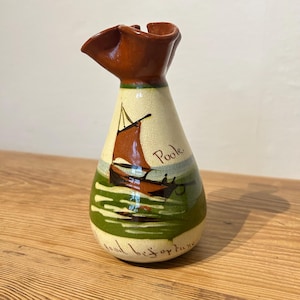 May include: A ceramic vase with a brown rim and a cream-coloured body. The vase features a painted sailboat scene with the word "Poole" and the phrase "good bes fortune". The base is green and the boat has a brown sail.