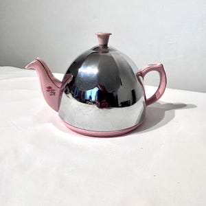 May include: A teapot featuring a polished chrome dome-shaped body with a pink base, handle, and spout. The spout is decorated with a small floral pattern of pink roses and green leaves. A matching pink lid with a small knob sits on top.