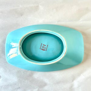May include: A vintage, rectangular, turquoise-colored dish with a central oval depression. The dish has a glossy finish and a white rim. The underside features a red and white stamp that reads "Wade England".