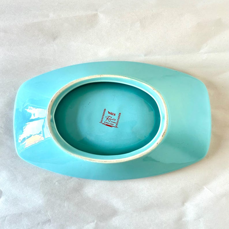 May include: A vintage, rectangular, turquoise-colored dish with a central oval depression. The dish has a glossy finish and a white rim. The underside features a red and white stamp that reads "Wade England".