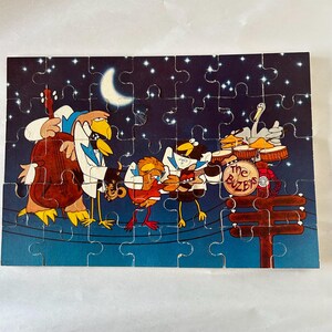 May include: A rectangular jigsaw puzzle featuring a cartoon band of birds called The Buzbys performing on a utility wire at night. The scene includes a crescent moon, stars, a double bass, a guitar, and a drum set with the text The Buzbys.