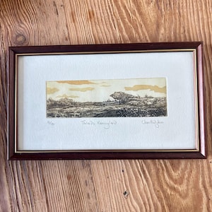 May include: A framed landscape print titled Towards Kessingland, numbered 31/150 and signed by the artist. The artwork features a horizontal etching of a field with a large tree under a yellow sky, set within a dark brown frame with gold trim.
