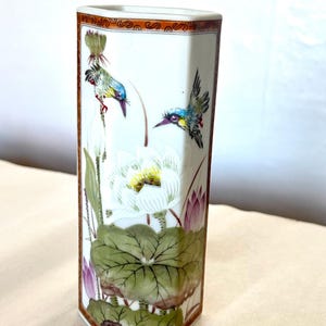 May include: A hexagonal ceramic vase featuring a white background with hand-painted illustrations of two colorful birds, white lotus flowers, and green lily pads. The top and bottom edges are decorated with an orange and black geometric border.