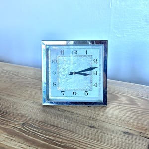 May include: A square, silver-toned clock with a light blue face and black hands. The clock face features black numerals and a rectangular inner border. The clock is set on a wooden surface.