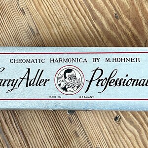May include: A rectangular silver-coloured case for a chromatic harmonica by M. Hohner. The lid features black text reading The Larry Adler Professional 16, a circular illustration of a person playing a harmonica, and the text Made in Germany.