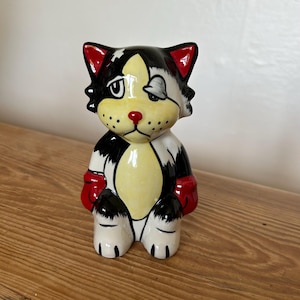May include: A ceramic figurine of a cartoon cat with black, white, and yellow markings. The cat has a red nose, red inner ears, and red boxing gloves. One eye is open and the other is covered by a grey patch. The figure has a glossy finish.