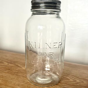May include: A clear glass jar with a ribbed metal screw-top lid. The front of the jar features embossed text that reads THE KILNER JAR IMPROVED REG. The glass is transparent, showing a cylindrical body with a wide mouth and a threaded neck.