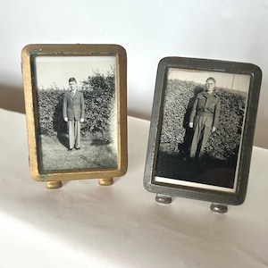 May include: Two antique metal photo frames, one gold and one silver, each containing a black and white photograph of a person standing before a hedge. The frames have a small, curved base for display on a table or shelf.