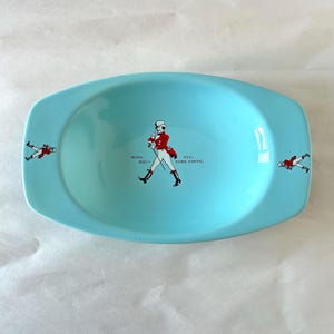 May include: A light blue oval ceramic dish featuring a central illustration of a man in a red coat and top hat, with the text "BORN 1820 - STILL GOING STRONG." Smaller versions of the same figure appear on the left and right sides of the dish.