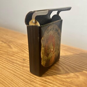 May include: A dark brown rectangular metal case with a hinged lid and a silver-toned metal latch mechanism. A circular sticker is applied to the front, featuring a reddish-brown illustration and the text "Zou je het wel doen?" in black.