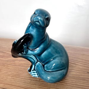May include: A teal ceramic sculpture of a seal with a black seal pup. The sculpture has a glossy finish and is sitting on a wooden surface. The seal is in a seated position, with the pup nestled in its arms. The sculpture is a decorative item.