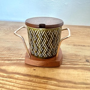 May include: A vintage ceramic cup with a wooden lid and base. The cup features a green and white geometric pattern and is held by a gold-colored metal handle. The wooden lid and base are a warm brown color.